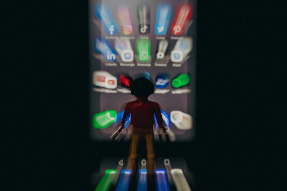 Toy figure facing a blurred phone screen of social app icons such as Facebook, Instagram and TikTok in the dark