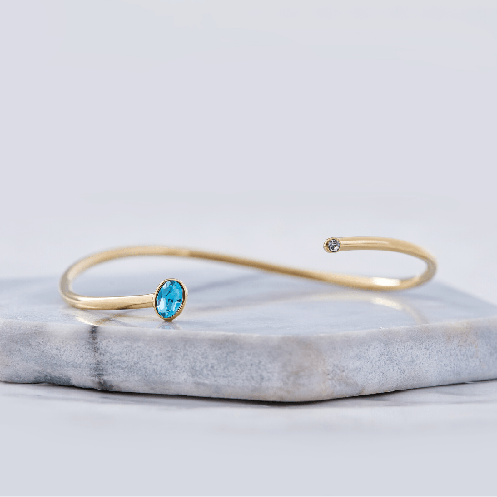 Gold open cuff bracelet set with a blue birthstone, resting on marble