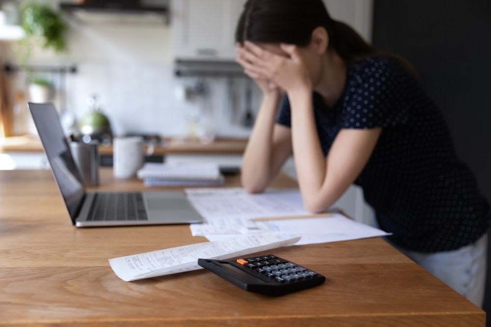 Woman with her head in her hands at a kitchen table covered in bills, with a calculator and a laptop