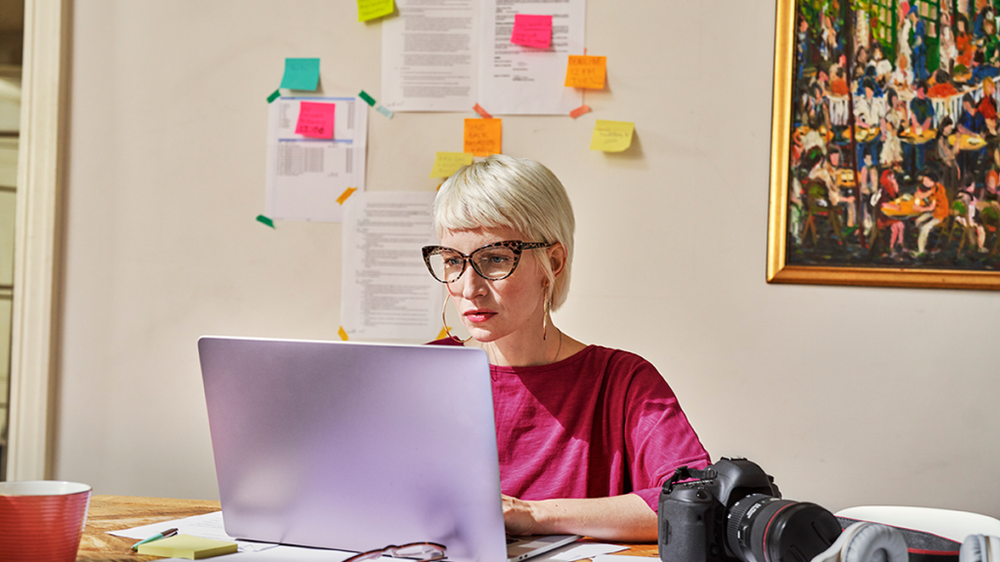 Woman in glasses working on a laptop at a desk, with a camera beside her and sticky notes on the wall