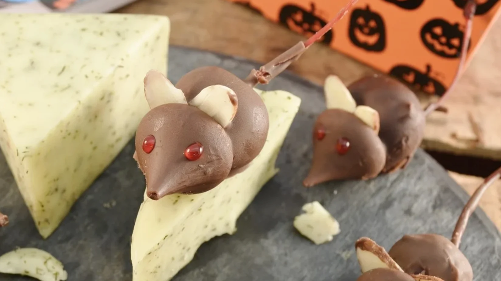 Chocolate mice with almond ears and red candy eyes on a wedge of herbed cheese, a Halloween box behind