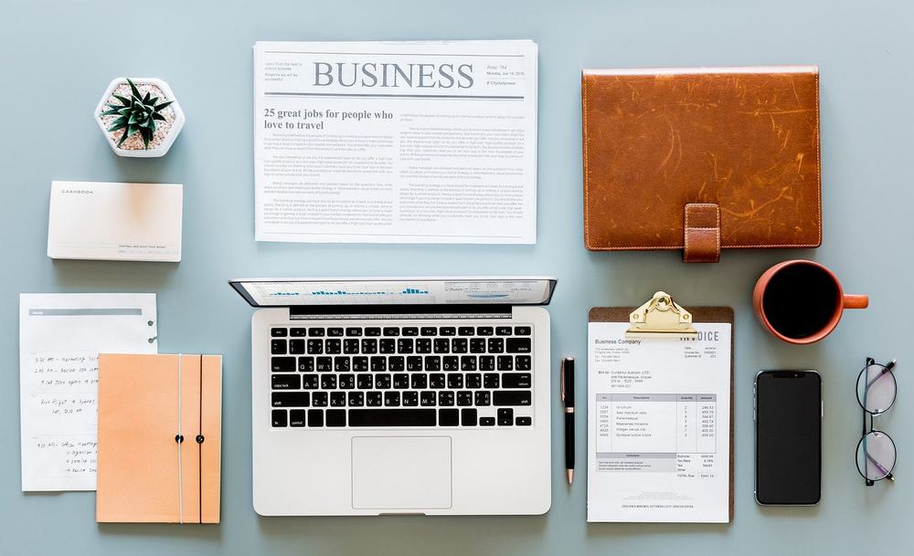 Neatly arranged desk from above: laptop, newspaper, leather planner, clipboard invoice, phone, glasses and coffee