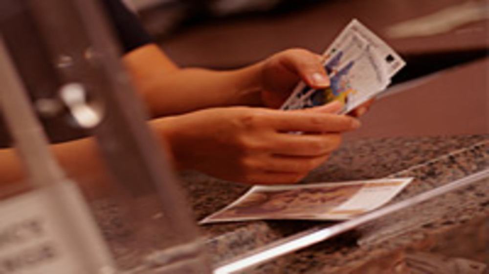 Hands holding banknotes at a bank teller's granite counter behind a glass screen