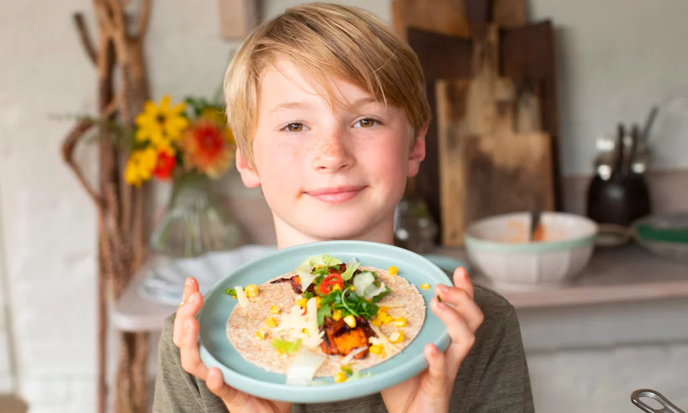 Smiling boy holding up a plate with a homemade tortilla topped with vegetables, corn and greens