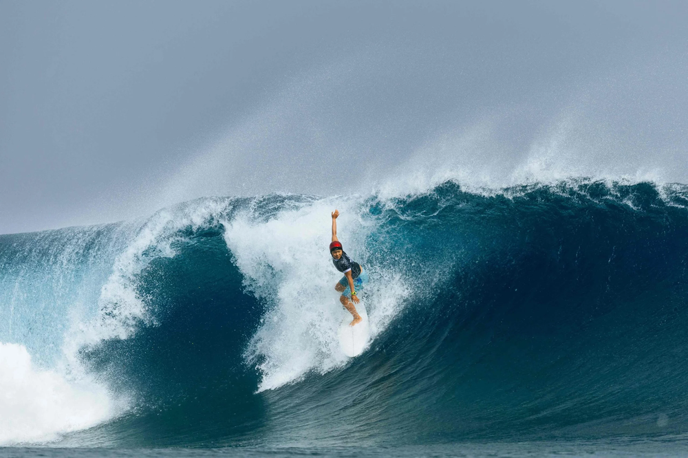 Surfer with one arm raised drops down the face of a huge curling wave under a stormy gray sky