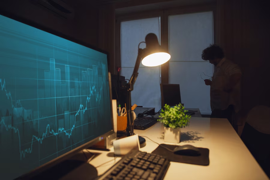 A dim office with a rising chart on a large monitor and a man by the window