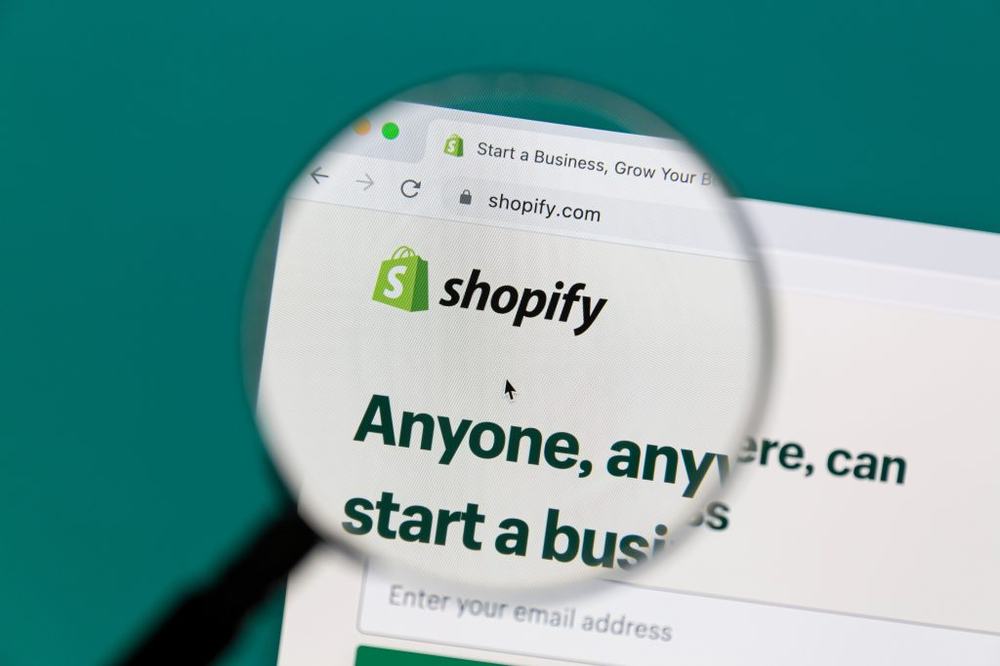 Magnifying glass over the Shopify homepage in a browser, reading 'Anyone, anywhere, can start a business'