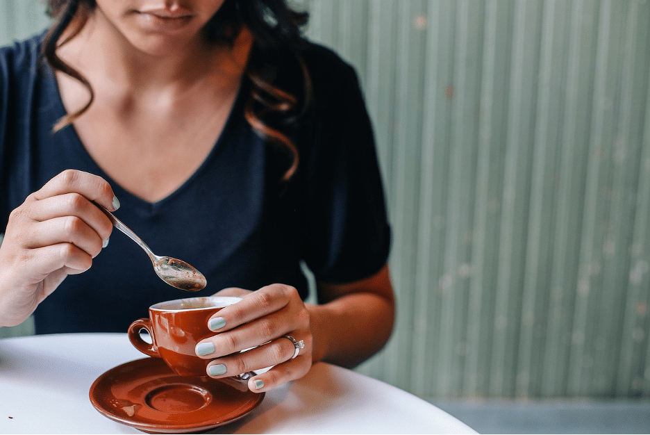 Woman lifting a spoon from a red cup of coffee on its saucer at a small white table