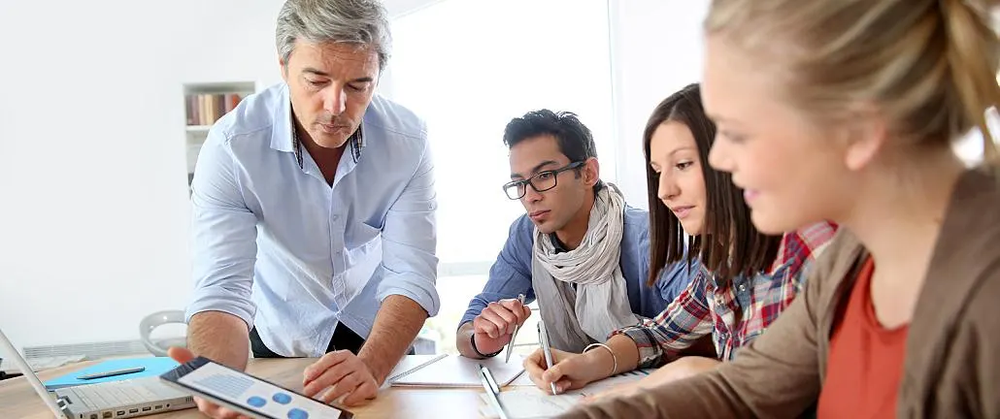 Mentor showing a tablet to three young colleagues taking notes around a table