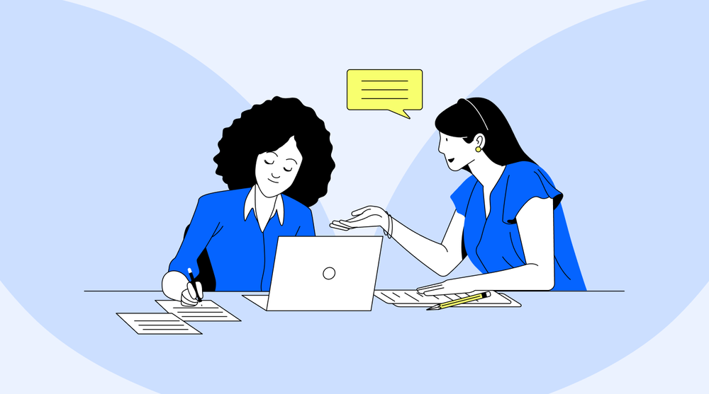 Two women at a desk with a laptop, one talking and gesturing while the other takes notes