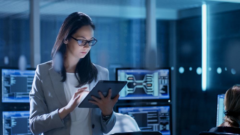 Woman in glasses reading a tablet in a dim control room lined with glowing monitors