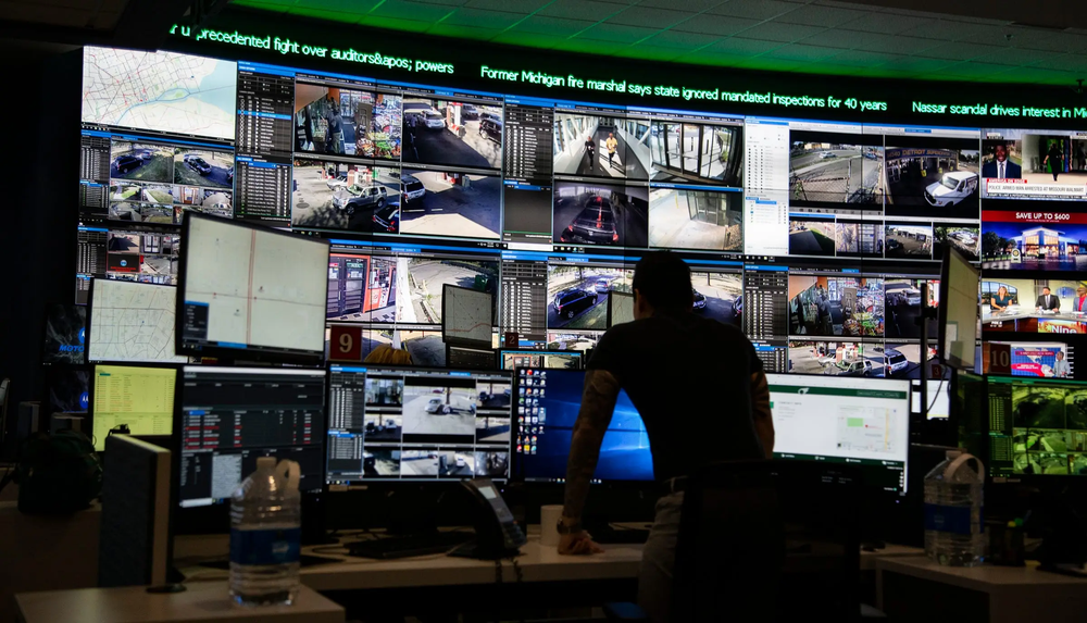 Man standing in a dark monitoring center before a wall of security camera feeds and news screens