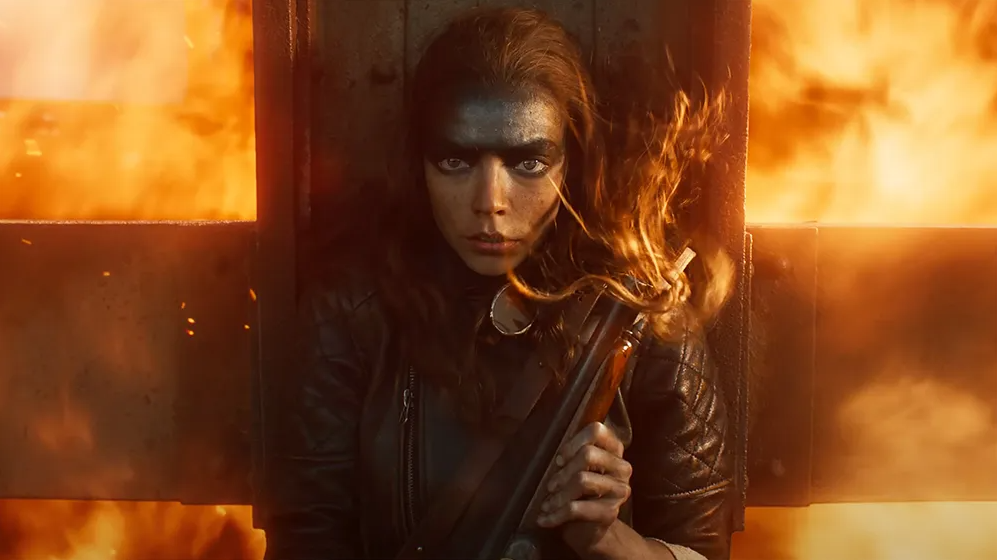 Furiosa, forehead smeared black, clutches a shotgun against a wall of flames in a still from the film