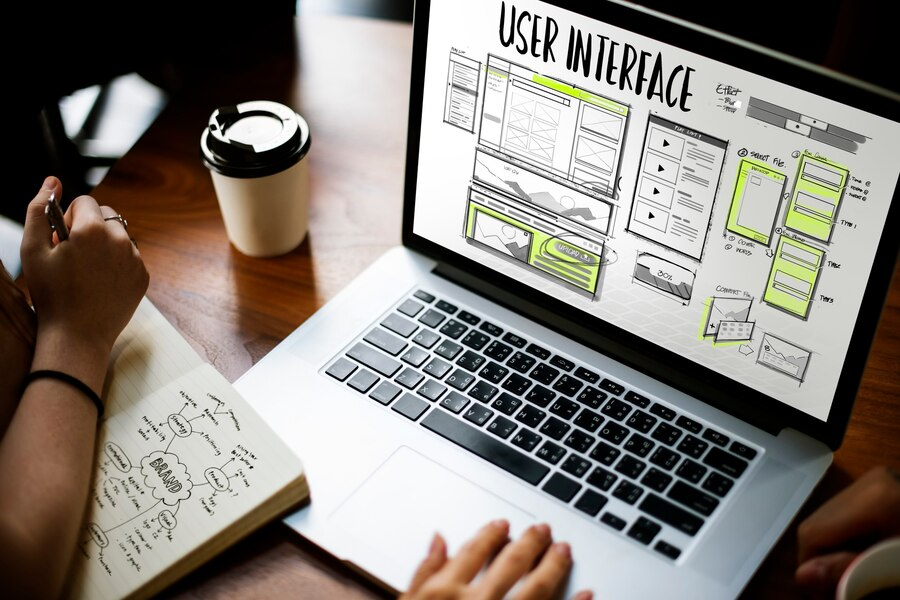 A laptop showing user interface wireframes beside a notebook of sketches and a coffee