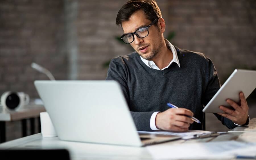 Man in glasses taking notes at a desk while reading a tablet, with an open laptop in front of him