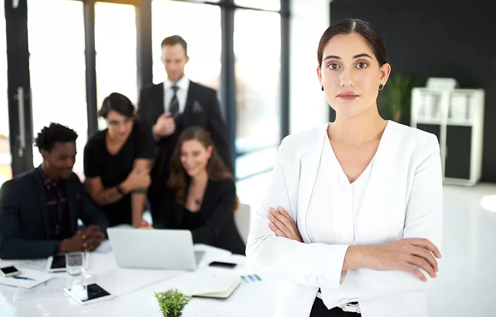 Woman with arms crossed stands apart while colleagues behind her laugh and talk around a laptop