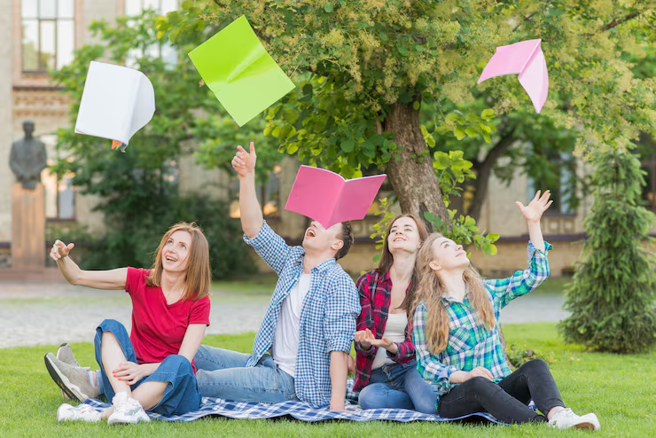 Four students sitting on a campus lawn, tossing notebooks into the air