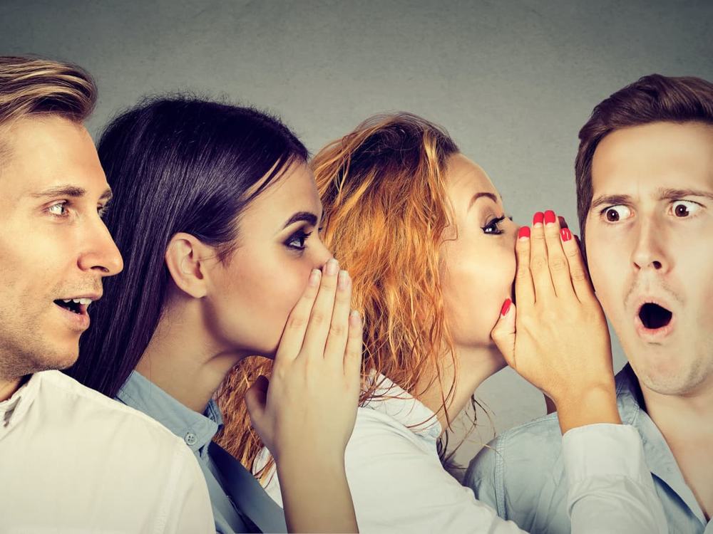 Four coworkers passing a whisper down a line, the last man wide-eyed and open-mouthed in shock