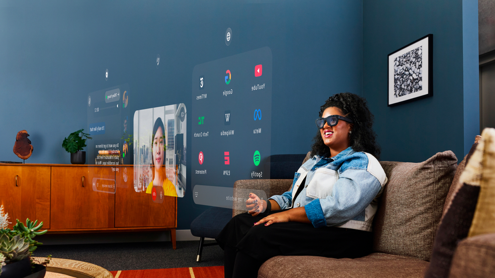 A woman on a sofa wearing mixed-reality glasses, with app windows and a video call floating beside her