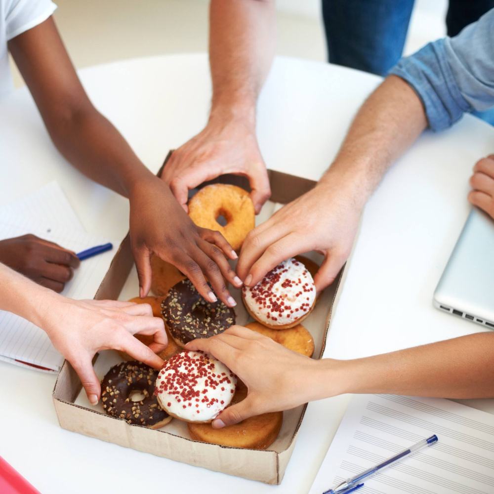 Several hands reaching into a box of glazed, chocolate and sprinkled doughnuts on a table