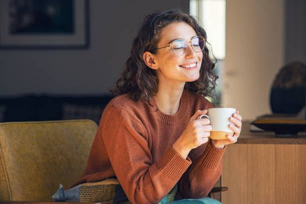Smiling woman in glasses holding a coffee mug with her eyes closed, relaxing by a window