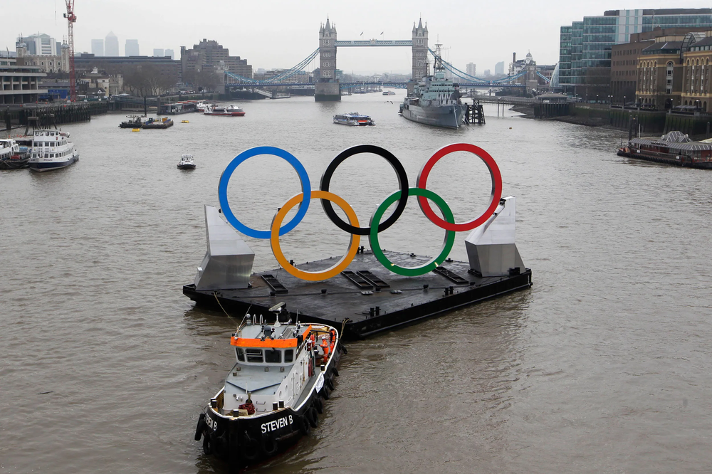 Giant Olympic rings on a barge towed along the River Thames by a tugboat, with Tower Bridge in the distance