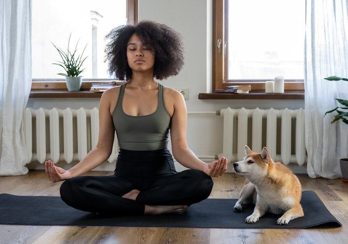 Woman meditating cross-legged on a yoga mat at home, eyes closed, with a Shiba Inu sitting beside her