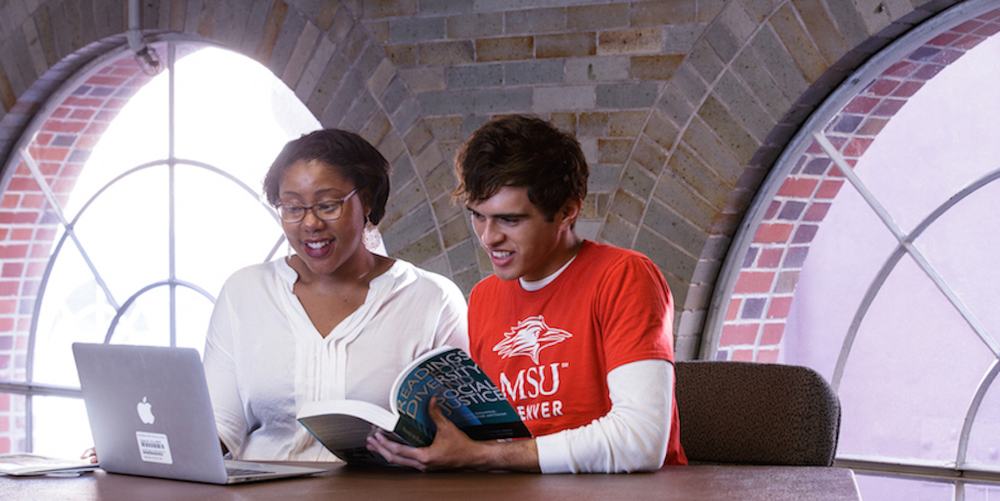 Two students reading a textbook together by an arched window, one in an MSU Denver T-shirt, a laptop beside them