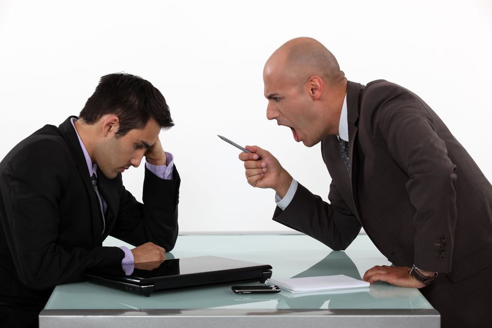 Angry boss shouting and jabbing a pen at an employee slumped over a closed laptop