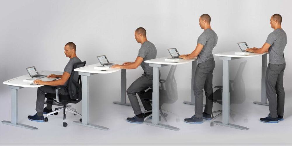 The same man at a sit-stand desk in four stages, rising from seated to standing as he types on a laptop