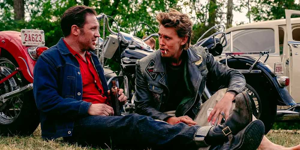 Tom Hardy and Austin Butler as bikers sitting on the grass beside their motorcycles in The Bikeriders