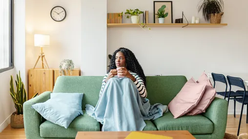 Woman wrapped in a blanket on a green sofa, holding a mug in a bright, plant-filled living room