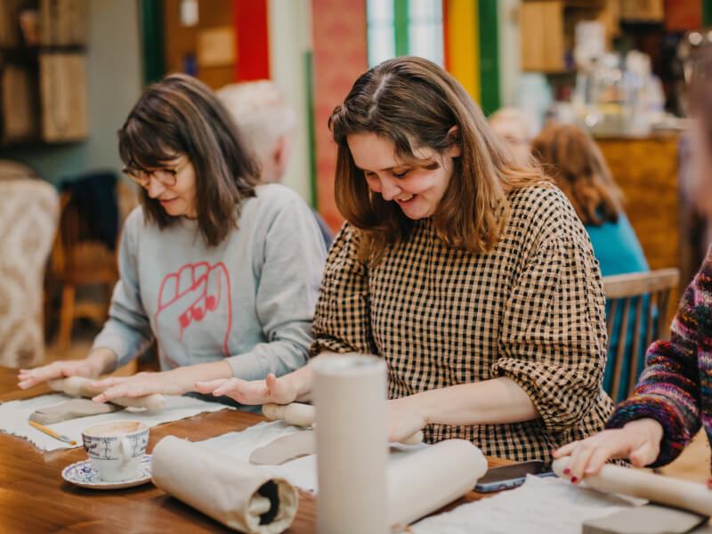 Two smiling women rolling clay at a table during a pottery workshop