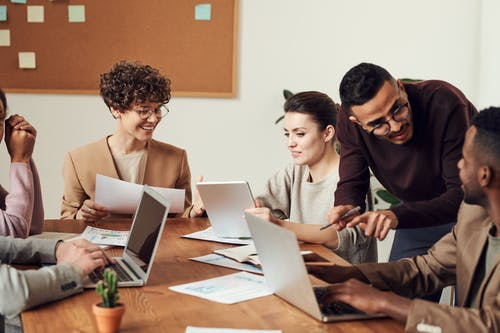 Coworkers smiling around a meeting table with laptops and papers as a colleague leans in to point at a document