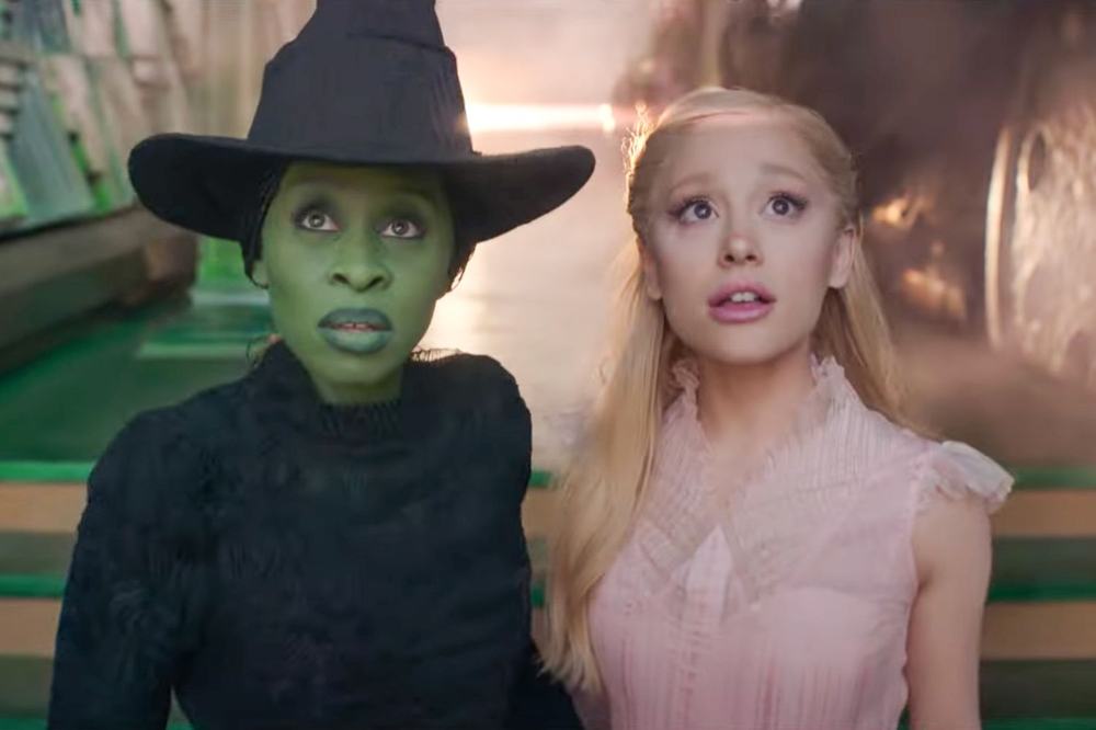 Elphaba, green-skinned in a black witch's hat, and Glinda, blonde in a pink dress, gaze upward in a still from Wicked